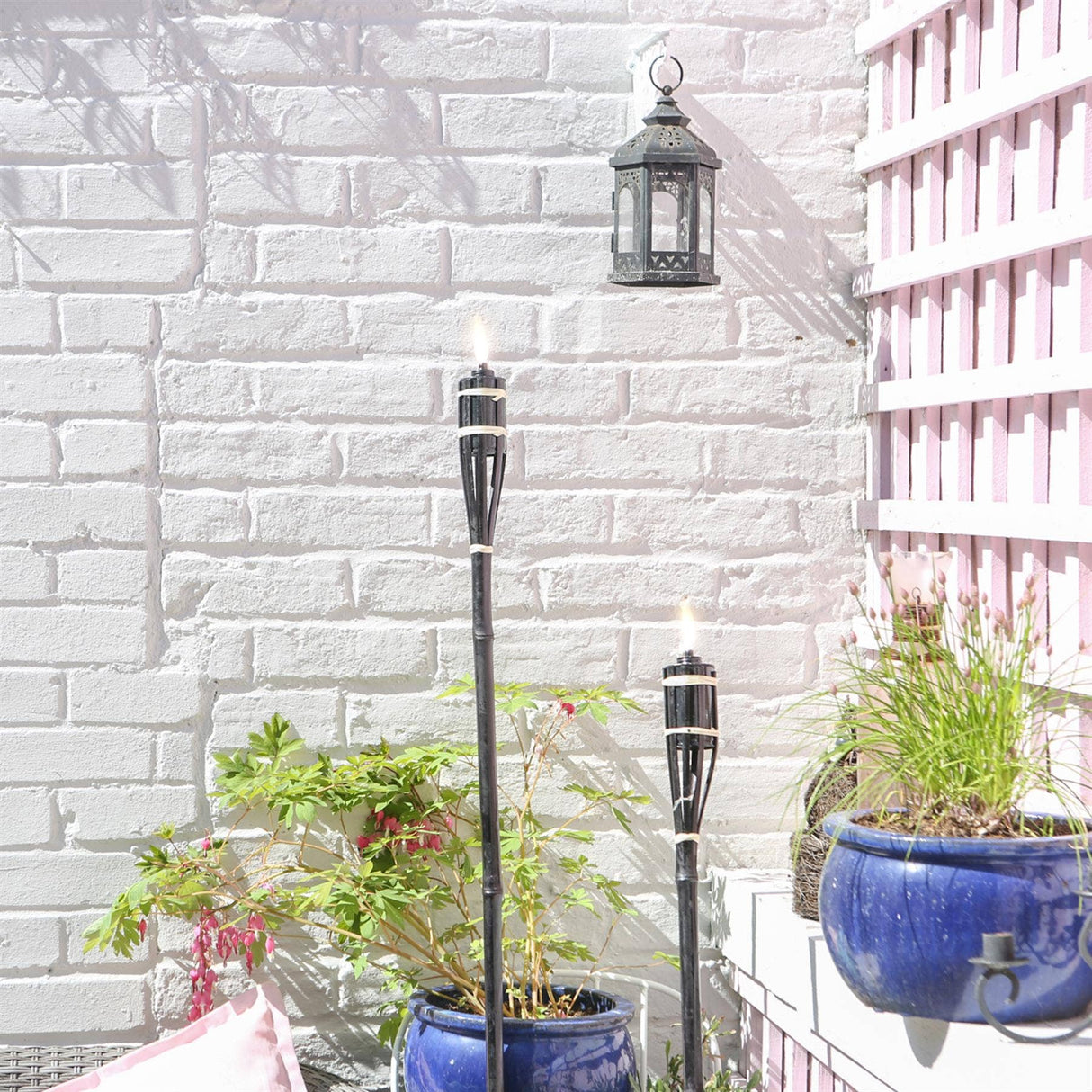 Tiki torches with lanterns on a white brick wall with plants and a blue pot.