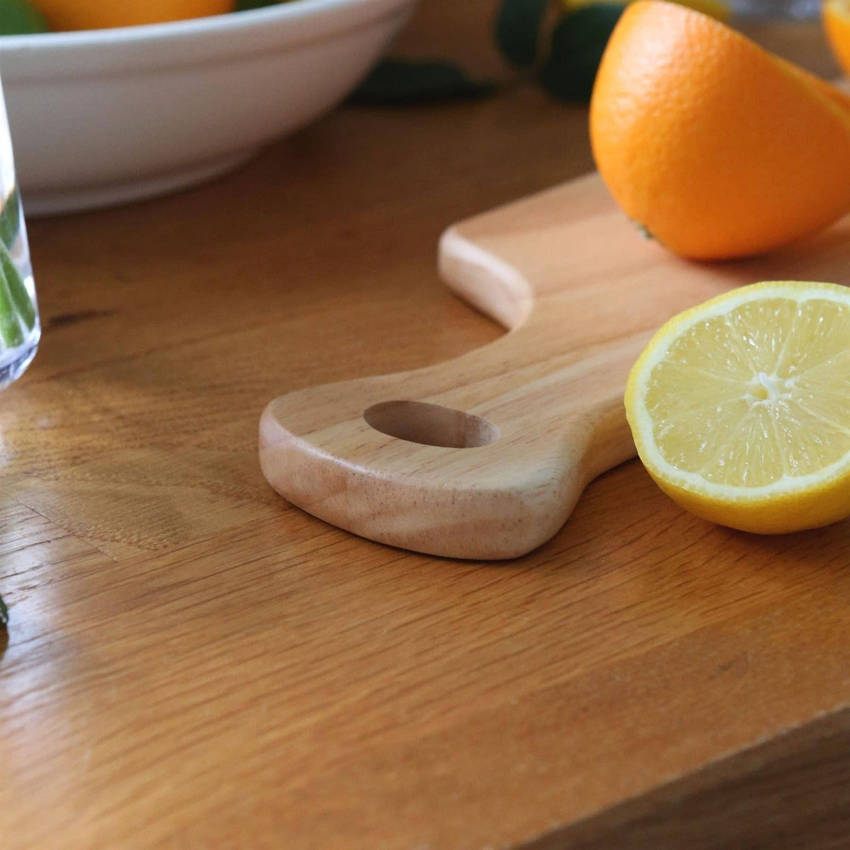 Close-up of the rounded wooden handle and hanging hole on the 34 × 16cm rubberwood board, beside a lemon half and sliced orange.
