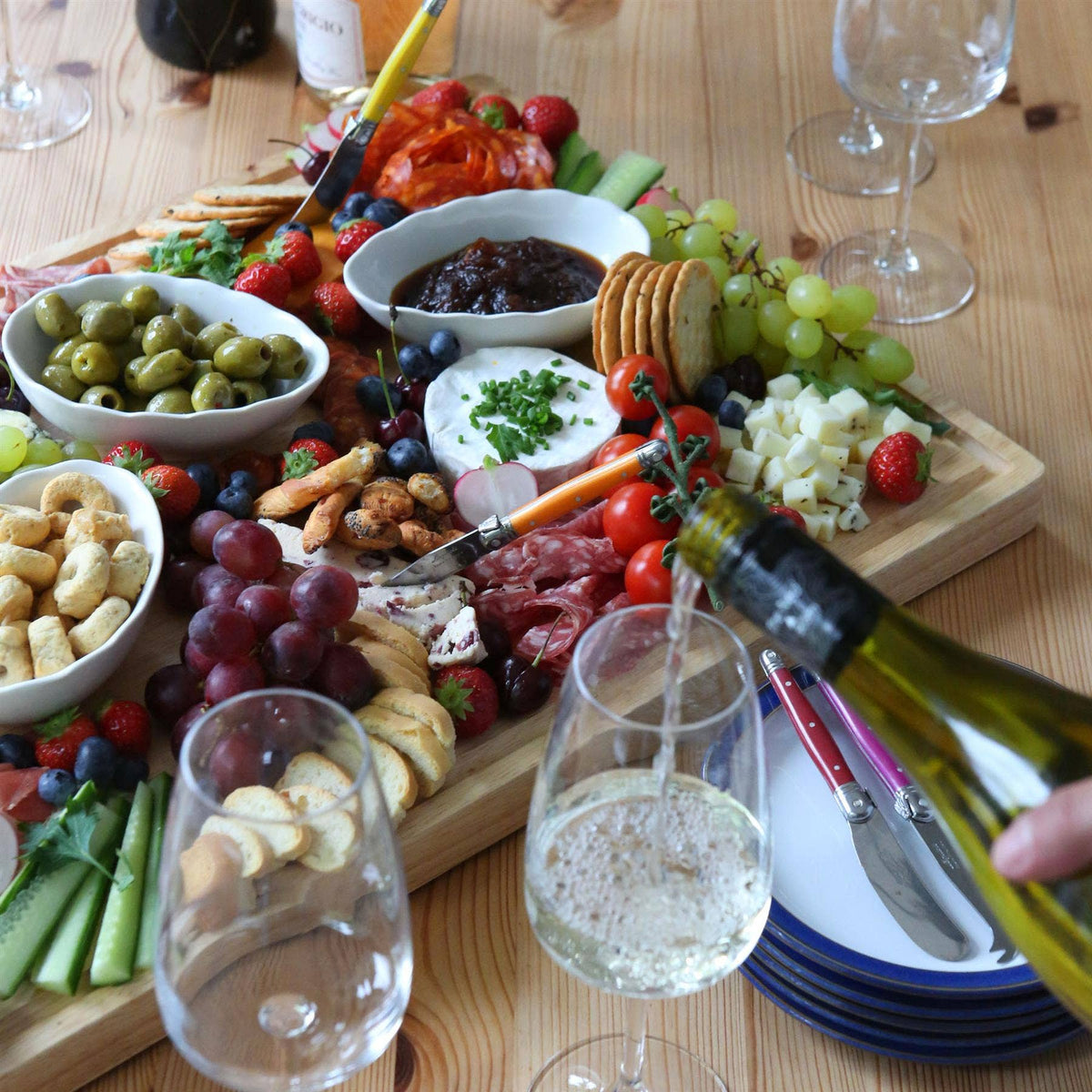 60 × 40cm rubberwood charcuterie board with cheeses, cured meats, crackers and fruit as white wine is poured beside it.