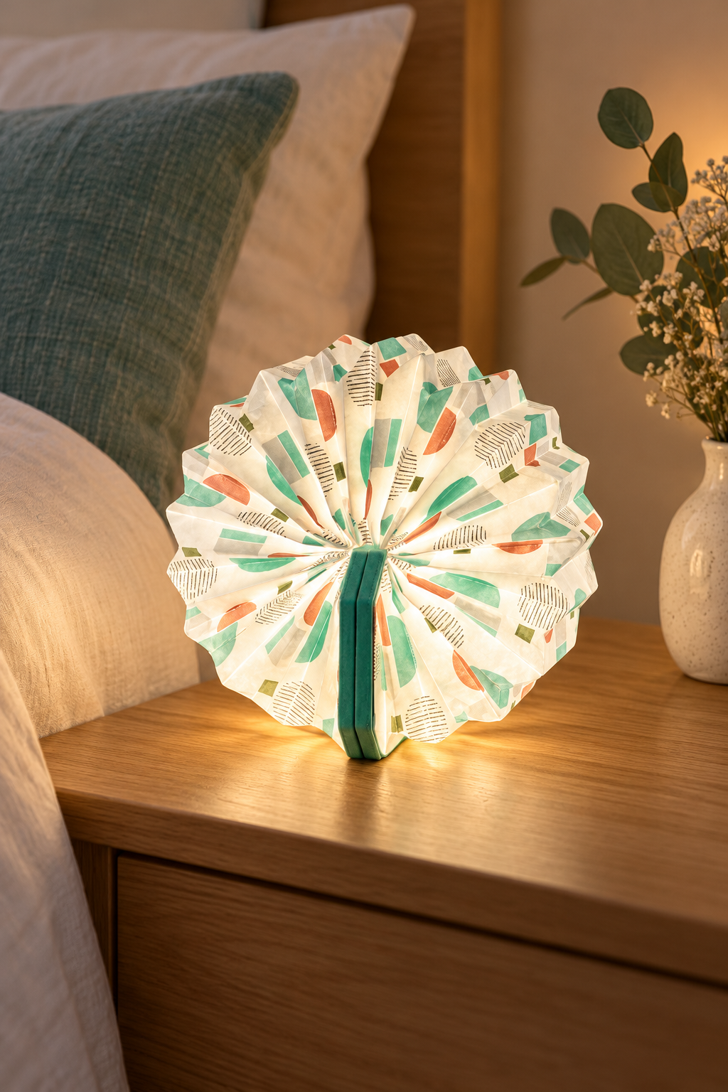Turquoise Geometric Velvet Accordion Lamp glowing on a bedside table beside cream bedding and a teal cushion.