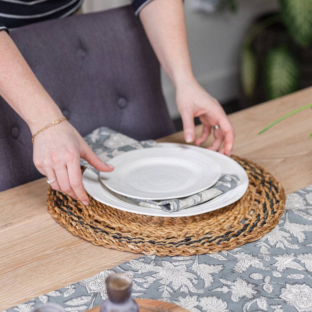 Hands placing a white plate on a woven placemat at a wooden dining table.