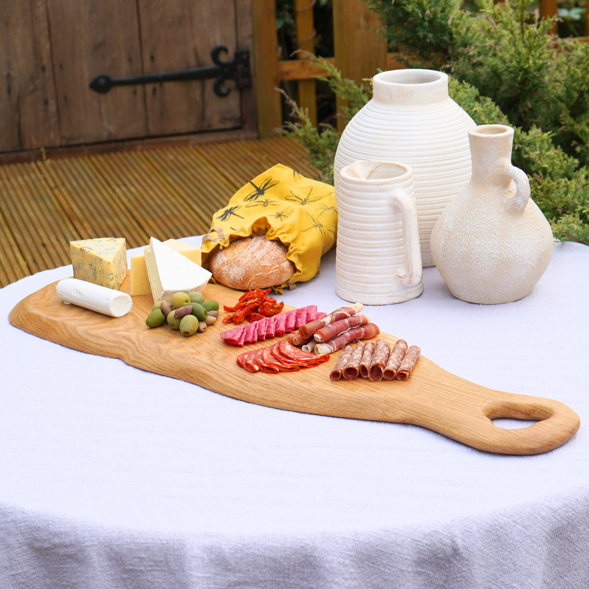 Wooden cutting board with charcuterie on a white tablecloth outdoors.