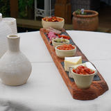 Dessert platter with small bowls of food on a wooden board, next to a textured ceramic vase on a white tablecloth.