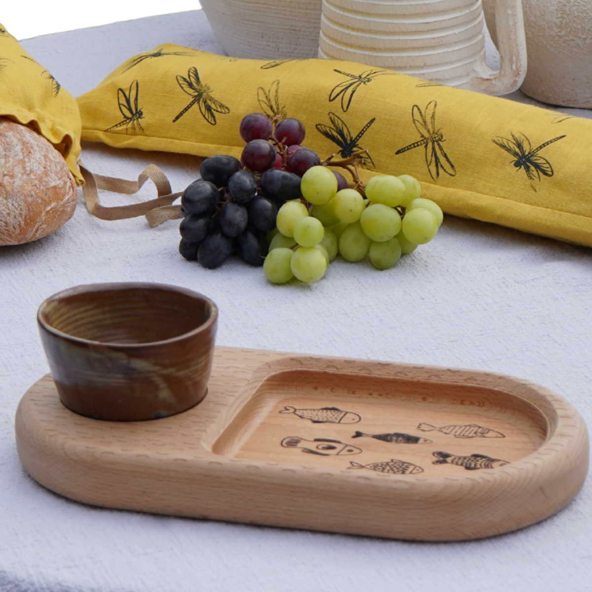 A wooden platter with a separate small bowl, grapes and a printed linen bread bag.