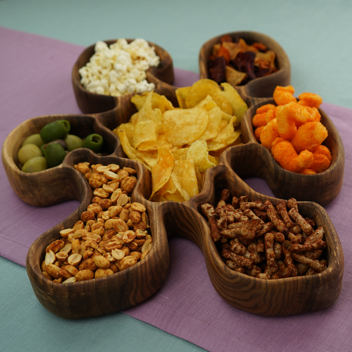 A divided wooden serving dish filled with crisps, nuts, olives and other snacks.