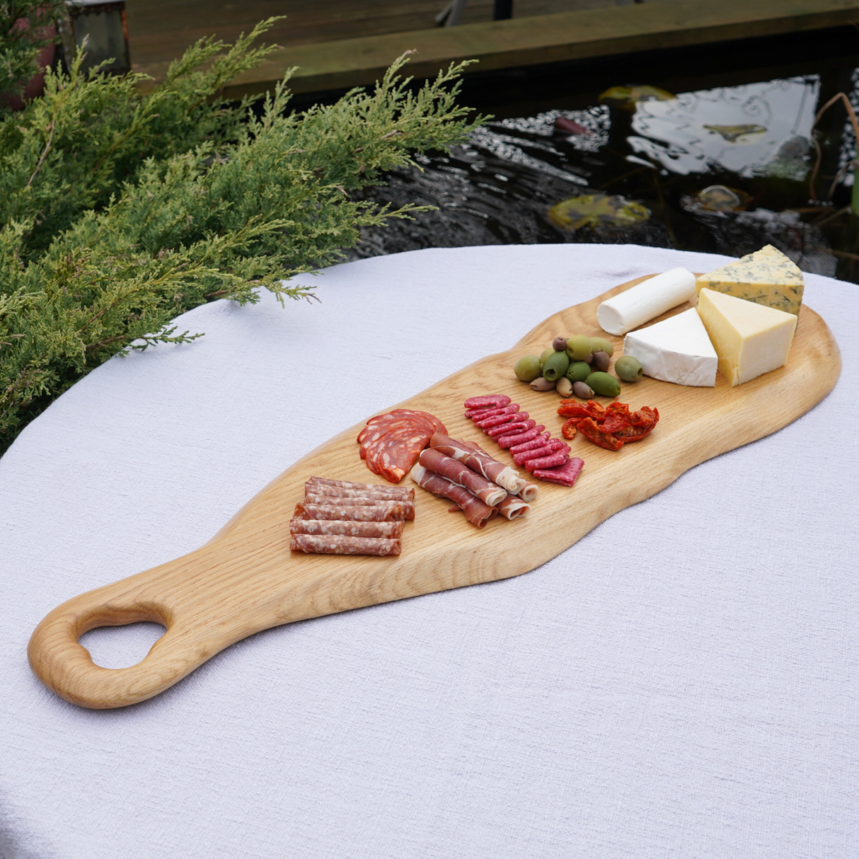 Wooden charcuterie board with meats, cheeses, and olives on a white surface outdoors.