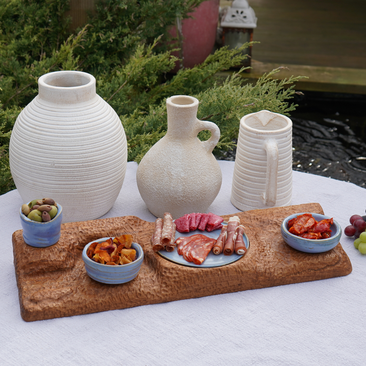 A wooden serving board with small bowls and food on an outdoor table beside white vases.