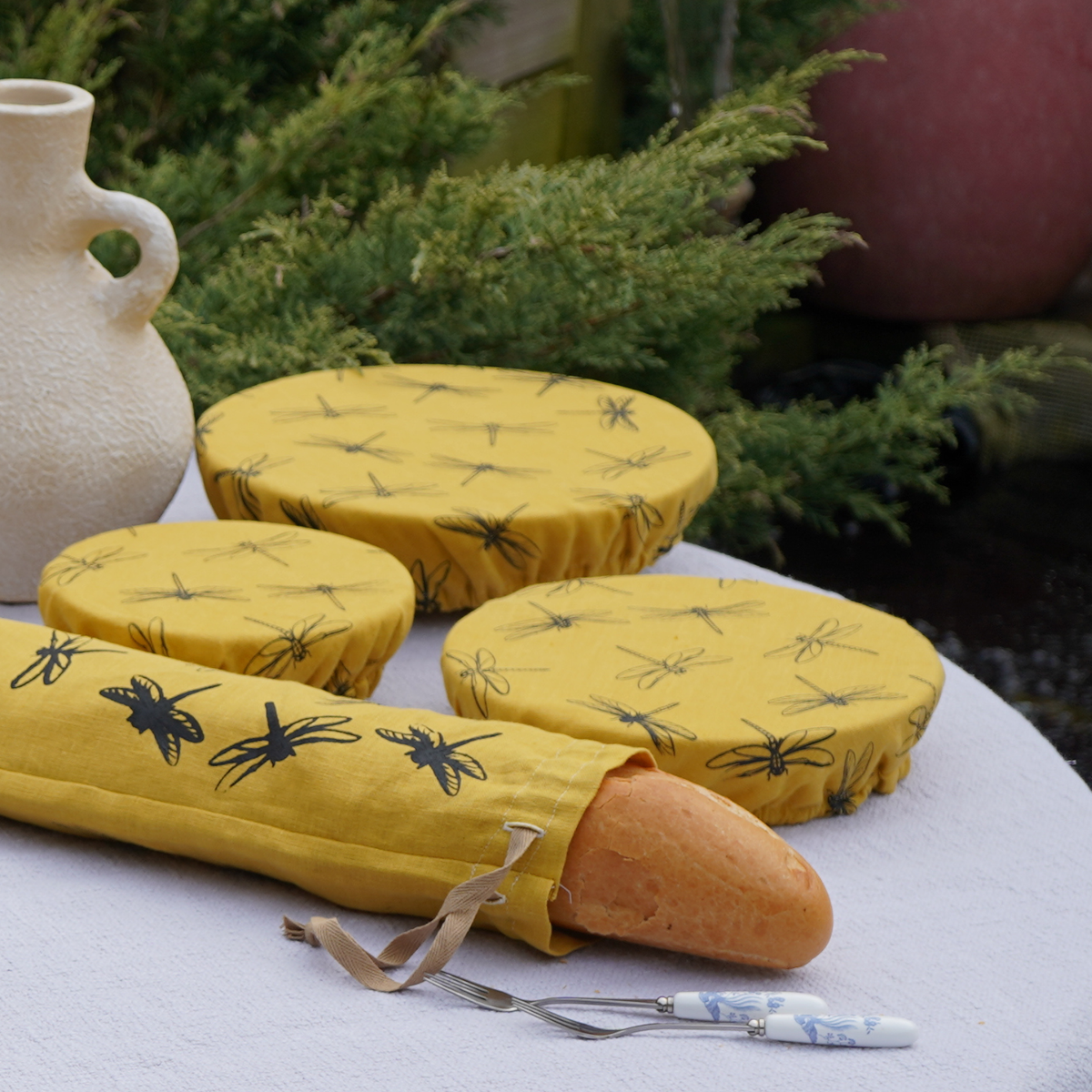 Yellow printed linen bowl covers and a matching bread bag beside fruit and a wooden spoon.