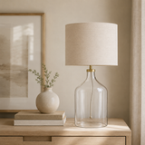 Table lamp with clear glass base and beige shade on a wooden surface, with a neutral wall and framed artwork in the background.
