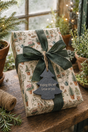 A wrapped gift box on a rustic wooden table, featuring cream wrapping paper with a vintage botanical pattern, a dark green ribbon bow, and a slate Christmas tree-shaped gift tag that reads 'Hoping this will Grow on you.' The scene is styled with potted plants, twine, and warm string lights in the background.