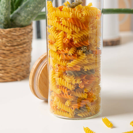 Glass container filled with multicolored rotini pasta on a white surface with a plant in the background.