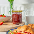 Jar of red jelly on a wooden board with a knife, surrounded by strawberries and croissants.