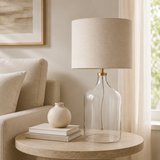 Decorative lamp with clear glass base and beige shade on a side table next to a sofa.