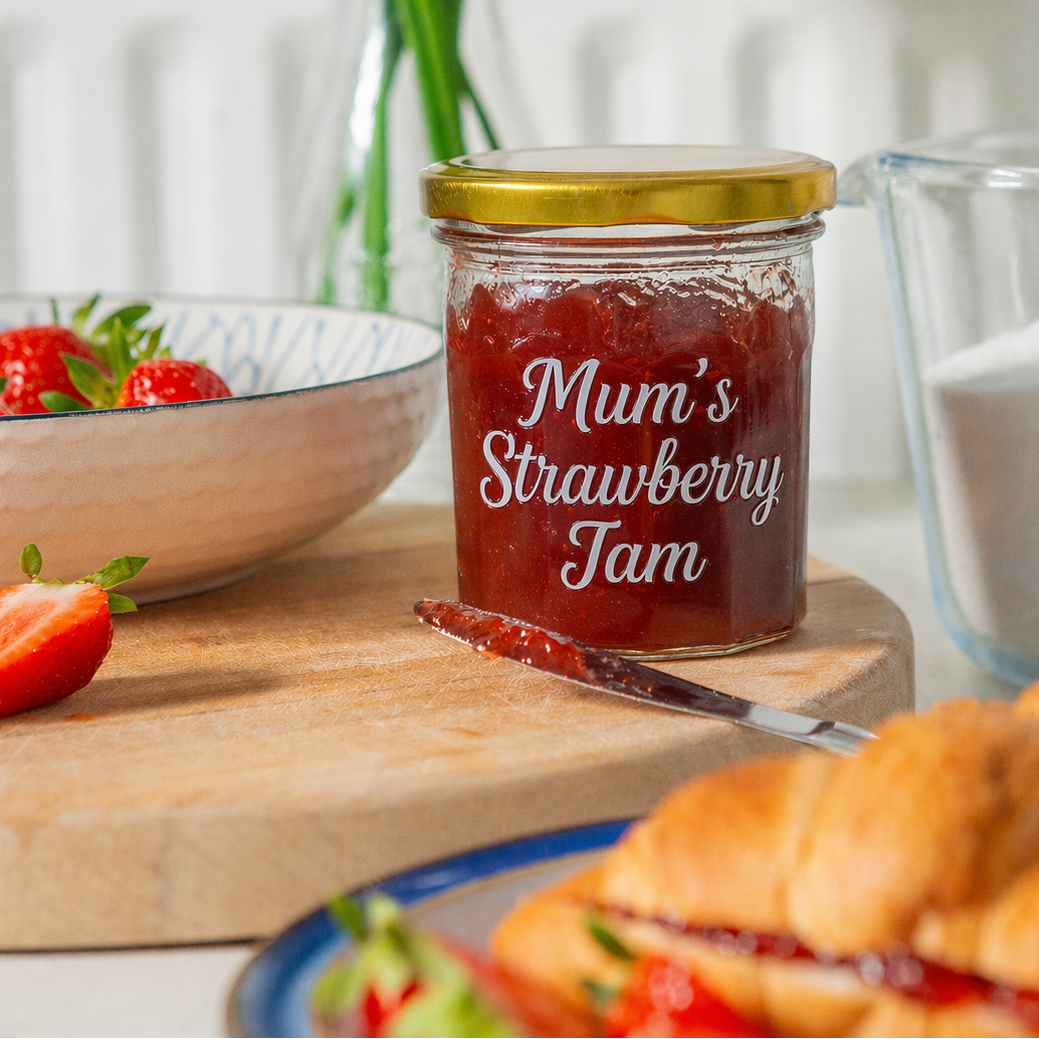 Jar of 'Mum's Strawberry Jam' on a wooden board with strawberries and croissants in the background.