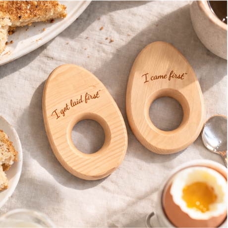 Two wooden egg-shaped coasters with humorous text on a breakfast setting.