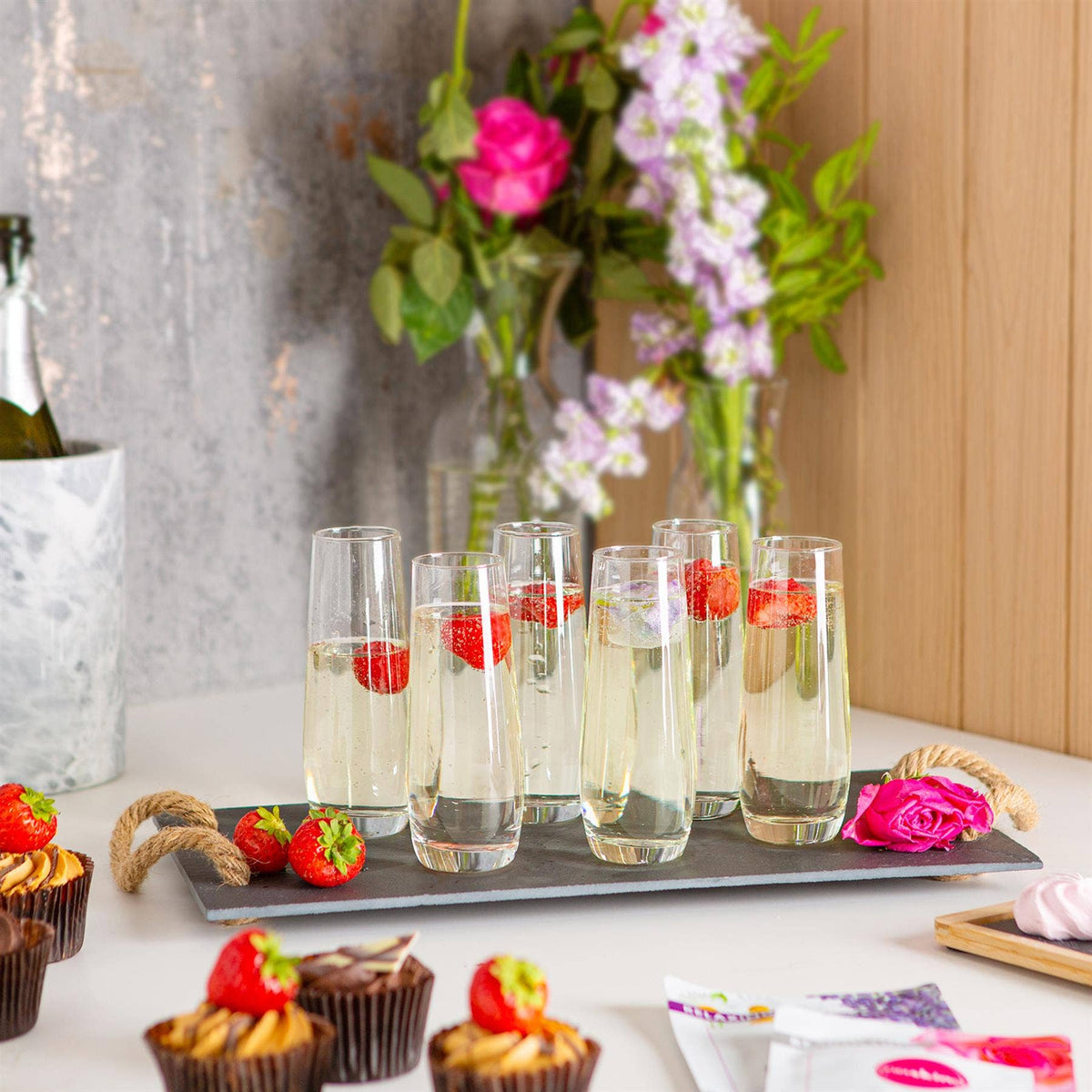 Clear champagne flutes with sparkling drinks and strawberries beside flowers and desserts.