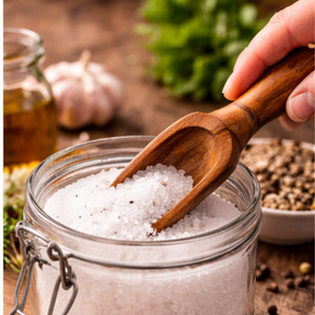 Wooden scoop taking salt from a glass jar on a wooden surface with herbs and spices.
