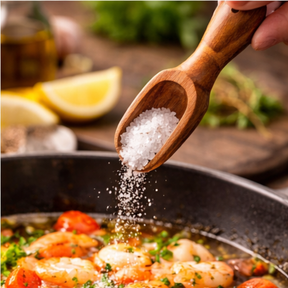 Shrimp being seasoned with salt from a wooden spoon in a pan.