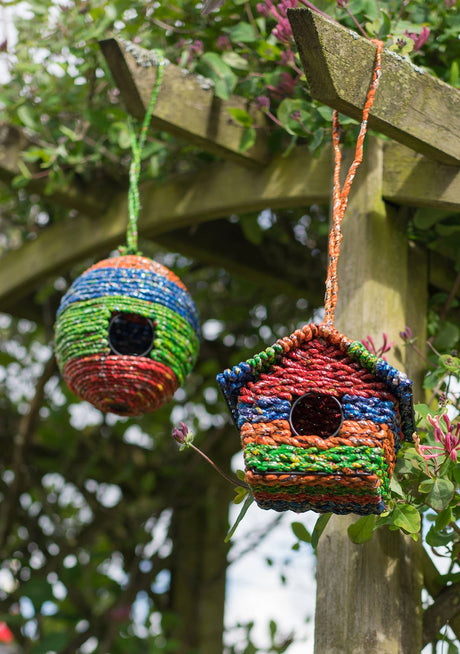 Colorful knitted birdhouses hanging from a wooden pergola with greenery in the background.