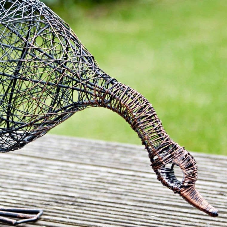 Wire sculpture of a duck drinking on a wooden surface with a blurred green background