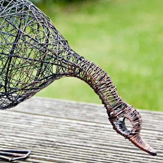 Wire sculpture of a duck drinking on a wooden surface with a blurred green background