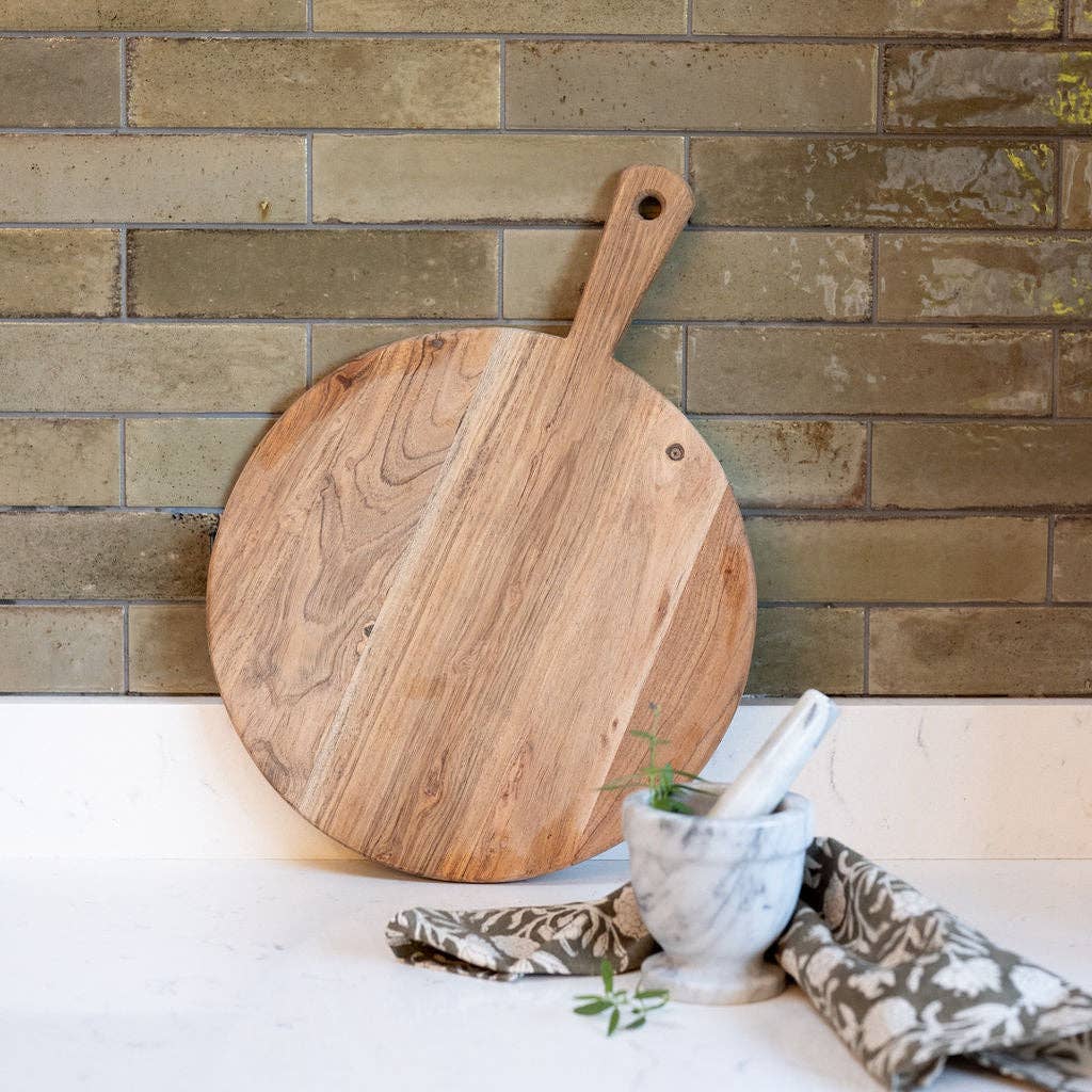 A round wooden serving board with a long handle standing on a kitchen counter.