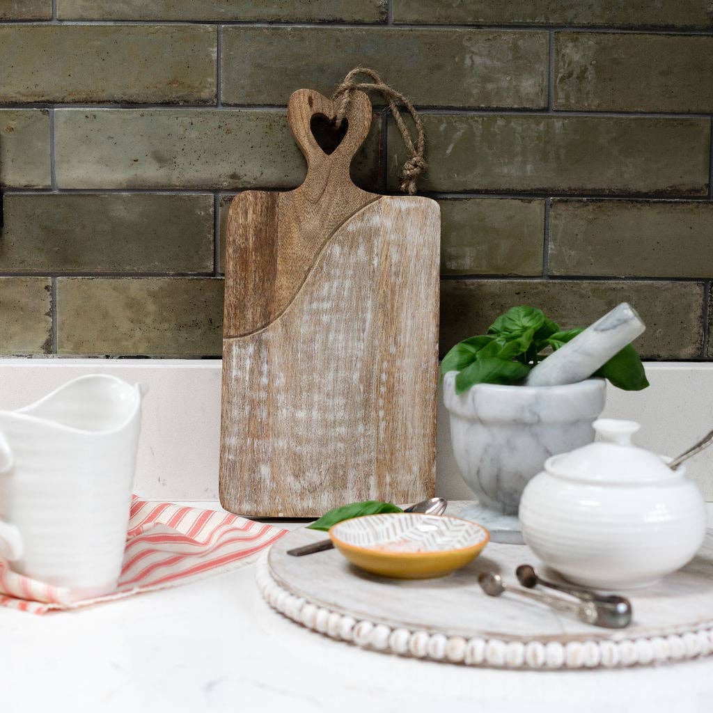 Wooden cutting board with heart handle on a kitchen counter with kitchen items.