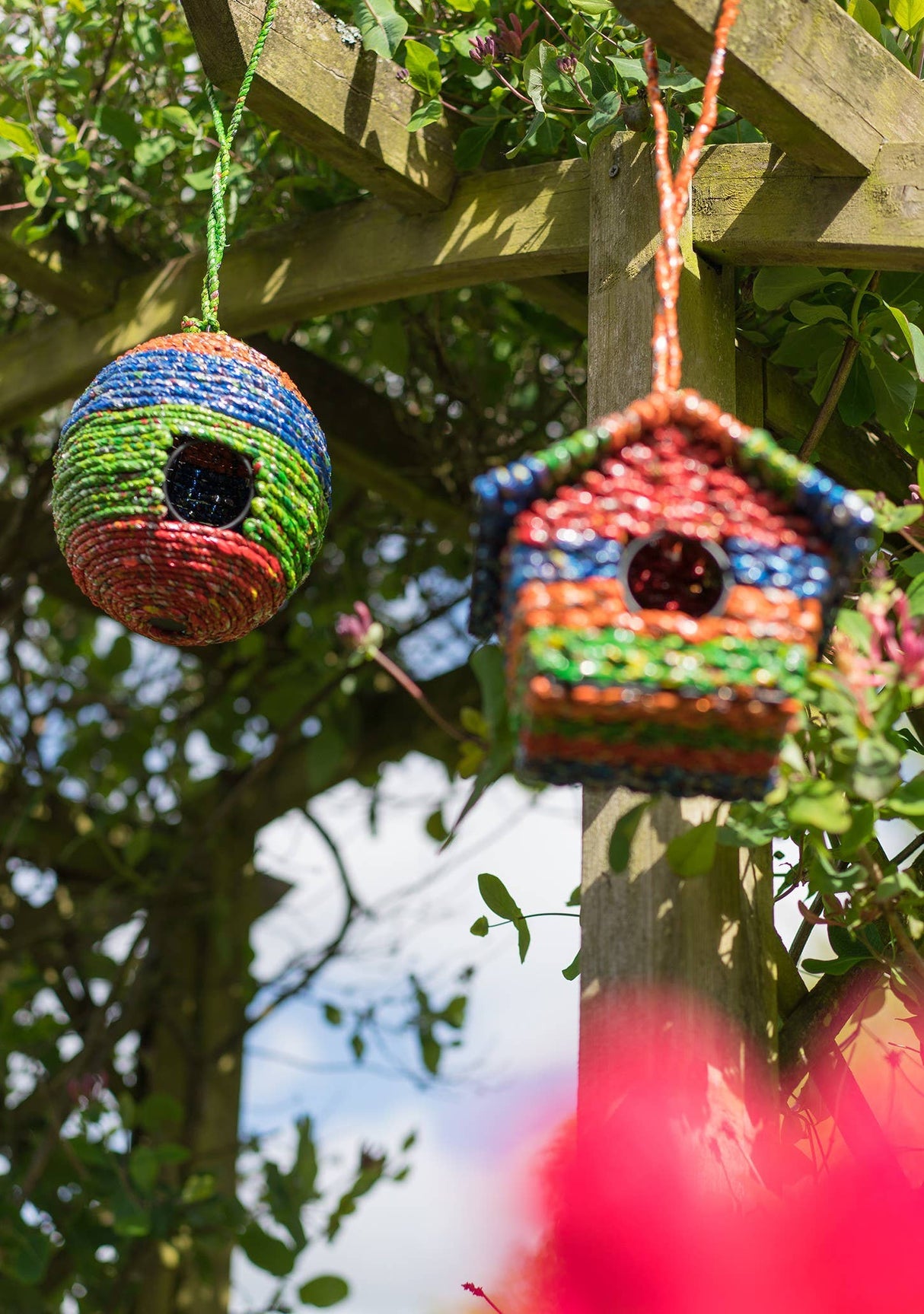 Colorful knitted birdhouses hanging from a wooden pergola with greenery in the background.