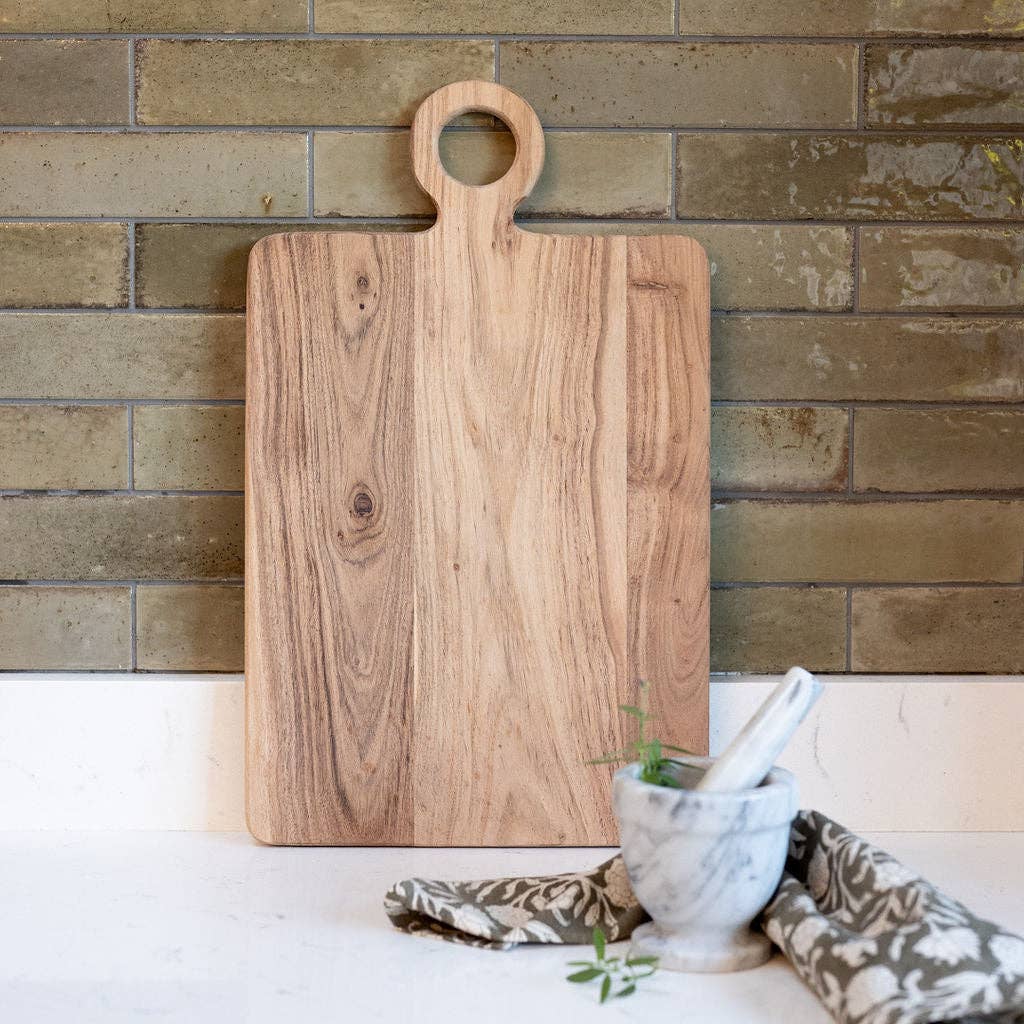 Wooden cutting board with a handle on a white surface against a brick wall.