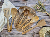 Set of wooden kitchen utensils on a rustic wooden surface with a woven mat and dried plants.