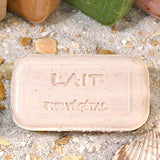 Bar of soap with 'Lait Therapeutique' branding on a sandy surface with seashells and plants.