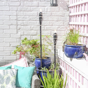 Outdoor patio with potted plants, torches, and colorful cushions against a white brick wall.