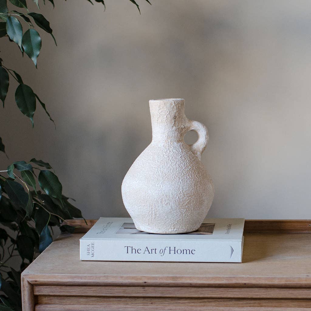 Textured white vase on a book titled 'The Art of Home' with a plant in the background