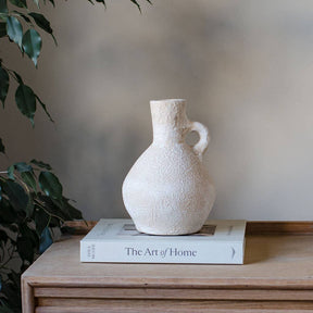 Textured white vase on a book titled 'The Art of Home' with a plant in the background