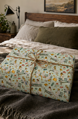 A rectangular gift box wrapped in a pale mint wrapping paper with a whimsical illustrated pattern of Ladies sitting on park benches, tied with natural twine, placed on a bed in a cozy bedroom setting.