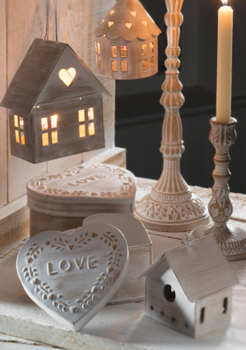 A collection of rustic, heart-shaped decorative home decor items including a wooden house lantern with a glowing heart window, a heart-shaped tin box, a small birdhouse ornament, and a candle holder, styled on a wooden surface.