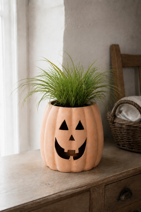 A decorative orange jack-o'-lantern planter with a carved face, filled with green ornamental grass, sitting on a rustic wooden dresser in a softly lit room.