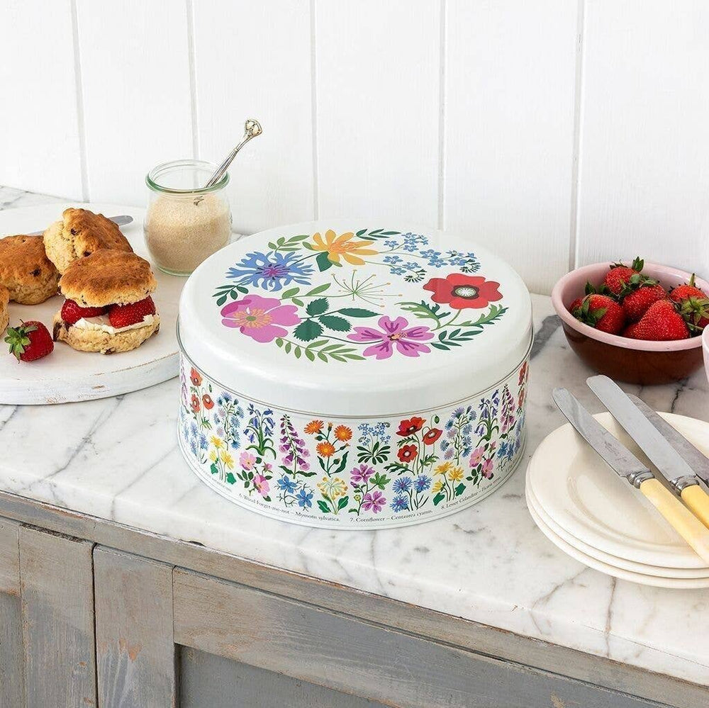 A round white tin with a colorful floral print sits on a marble kitchen counter beside scones, strawberries, and plates.