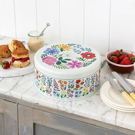 A round white tin with a colorful floral print sits on a marble kitchen counter beside scones, strawberries, and plates.