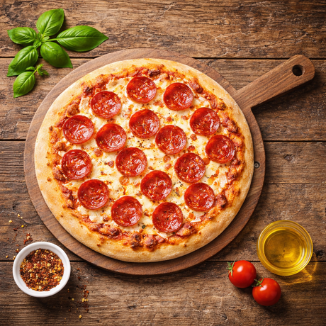 Pepperoni pizza on a wooden board with basil, tomatoes, and olive oil on a rustic wooden table.