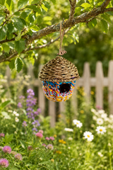 Colorful woven birdhouse hanging from a tree branch in a garden with flowers and a fence in the background.