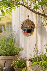 Colorful woven birdhouse hanging from a tree branch in a garden setting.