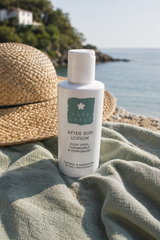A bottle of Pure Lakes After Sun Lotion stands on a textured sage green cloth next to a woven straw sun hat, with a sunny coastal beach and sea in the background.