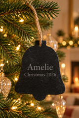 A black slate Christmas bell ornament engraved with 'Amelie Christmas 2026', hanging from a green pine tree branch with warm string lights in the background.