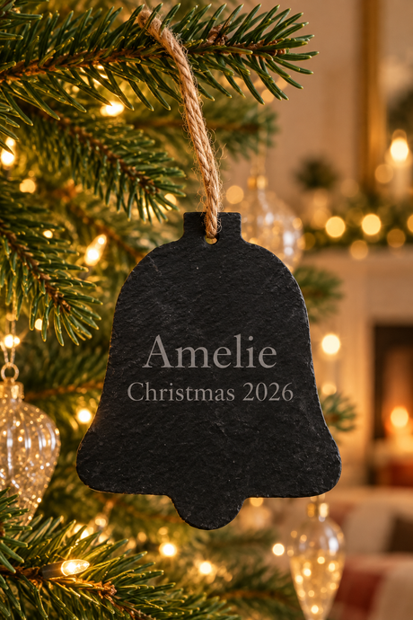 A black slate Christmas bell ornament engraved with 'Amelie Christmas 2026', hanging from a green pine tree branch with warm string lights in the background.