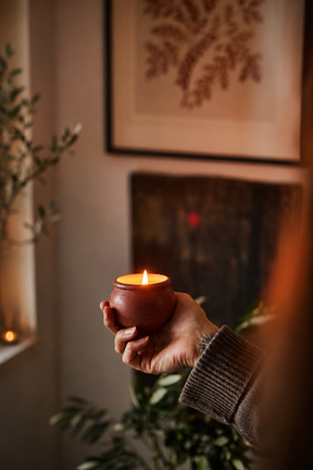 Hand holding a small lit terracotta pot candle in a cozy indoor setting with plants and framed artwork.