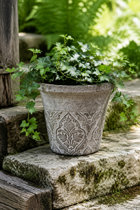 A gray stone-look planter with an embossed floral pattern, filled with green ivy plants, set on rustic stone steps outdoors with blurred green foliage in the background.