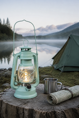 A mint green vintage-style lantern with a glowing light inside sits on a tree stump beside a metal cup and rolled map, with a camping tent, lake, and misty mountains in the background.