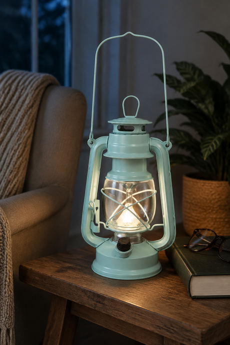 A vintage-style teal lantern lamp with a glowing light inside, placed on a wooden table in a cozy living room setting next to a chair and a potted plant.