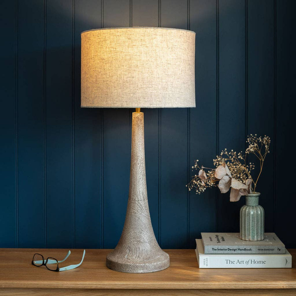 A table lamp with a textured, tapered stone-like base and a beige linen drum shade, lit up and placed on a wooden table against a dark blue paneled wall. A pair of eyeglasses, a small vase with dried flowers, and a stack of books are nearby.